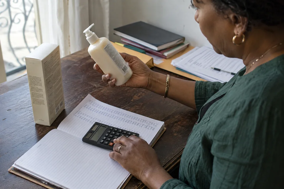 Une responsable commerciale calcule le prix d’un produit avec une calculatrice, des documents et un flacon sur son bureau.