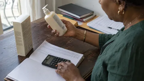 Une responsable commerciale calcule le prix d’un produit avec une calculatrice, des documents et un flacon sur son bureau.