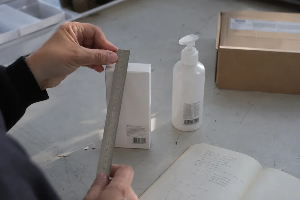 Des mains mesurent un étui de produit avec une règle métallique sur une table de préparation.