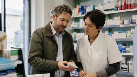 A sales rep shows a skincare sample to a pharmacist at the counter.