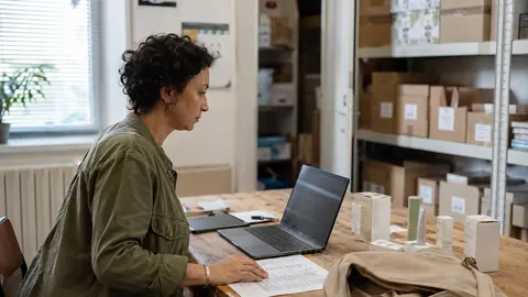 Une commerciale prépare sa visite sur un ordinateur, avec une fiche client, des échantillons et sa sacoche sur une table de travail.