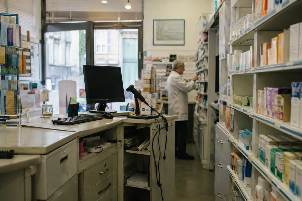 The counter and shelving of a neighbourhood pharmacy, with a pharmacist in the background.