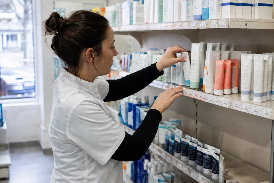A member of staff tidies skincare tubes on a pharmacy shelf.