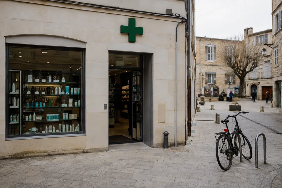 Une pharmacie de centre-ville, croix verte au-dessus de la vitrine, sur une place pavée où un vélo est attaché.