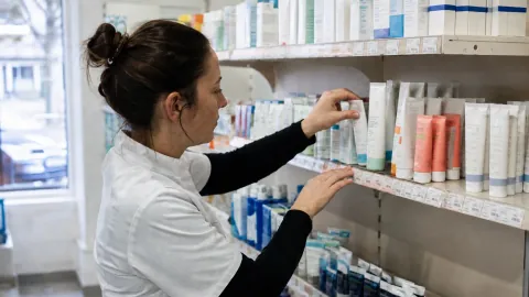 Une employée en blouse blanche range des tubes de soins sur les étagères d'une pharmacie.