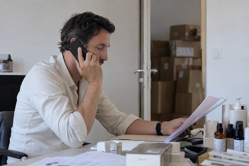 Un responsable de marque téléphone à son bureau en consultant un dossier, près de quelques produits de soin.