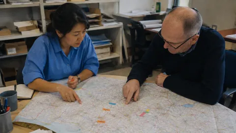 Two sales managers study a road map spread out on an office table.